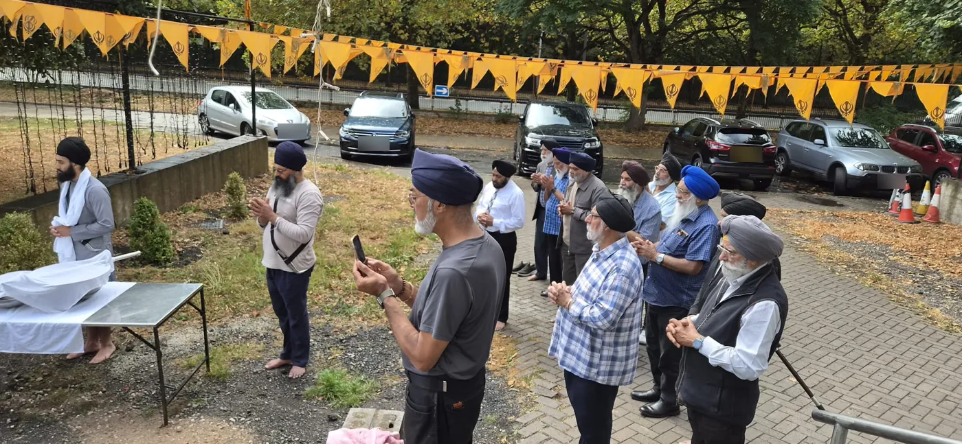 Members of the sangat standing with folded hands in Ardas beside the Nishan Sahib site, with Khanda bunting overhead