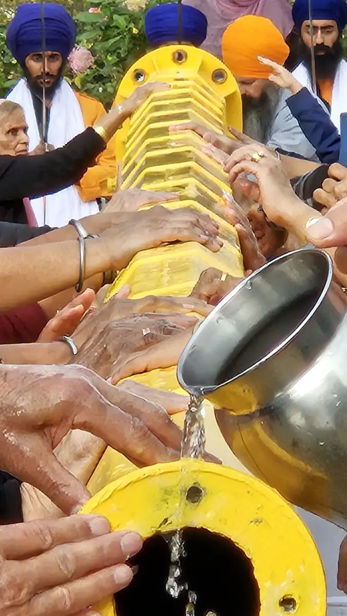 Water pouring from a steel jug over the yellow Nishan Sahib pole held by many hands